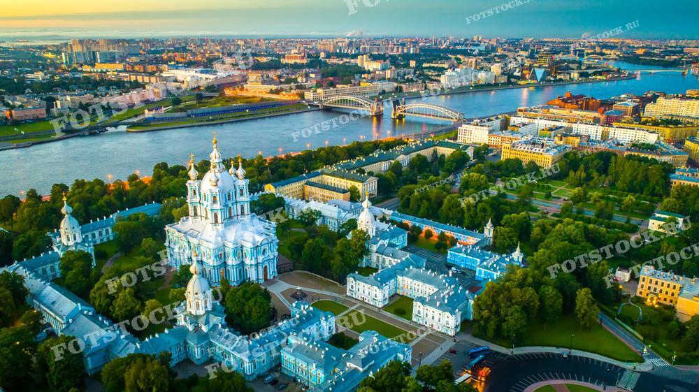 Фотография с высоты птичьего полёта показывает Смольный собор в Санкт-Петербурге, окружённый зелёными насаждениями Смольного парка. На заднем плане простираются река Нева и городские мосты, соединяющие берега, а вдали видны современные и исторические районы города. Игра света и теней придаёт этой сцене живописность и глубину, делая снимок идеальным для культурных публикаций, туристических брошюр и как эстетически приятный элемент декора.